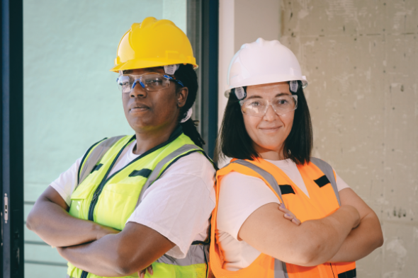 Two women stand in construction safety gear smiling with their arms crossed