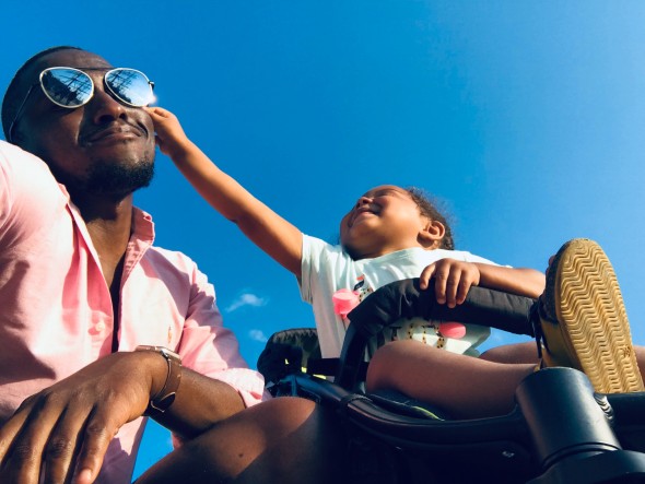 Man in sunglasses kneels next to child trying to grab the sunglasses with a blue sky background