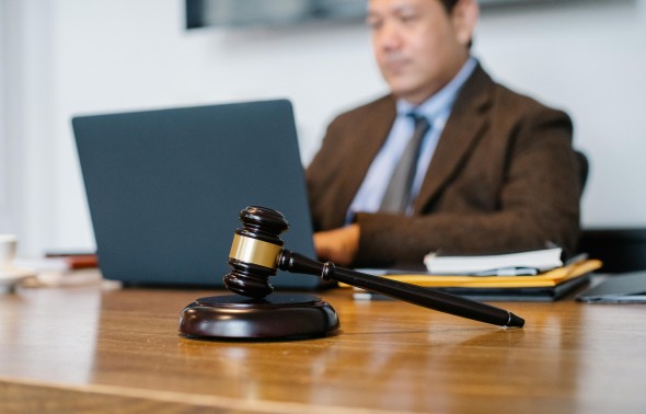 Gavel sits on a desk in the foreground with a man in a suit sitting behind the desk in the background
